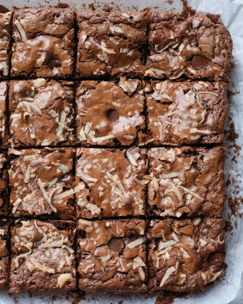 A close-up of a single fudgy coconut brownie with a glossy chocolate top and desiccated coconut, placed on a white marble surface with tropical leaves in the background.